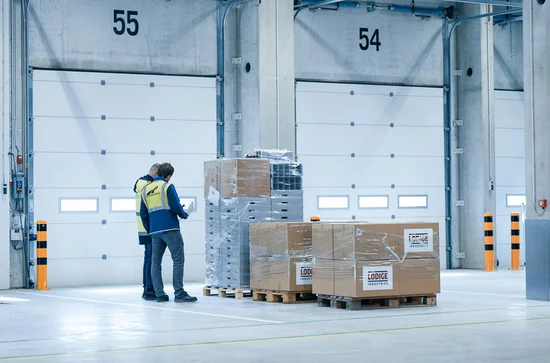 Two logistics staff checking shipment pallets in front of closed truck dock doors inside an air cargo terminal, ensuring accurate documentation and loading preparation.